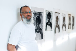 Image of artist Meleko Mokogosi looking toward the camera while leaning up against a wall in a gallery of his work.