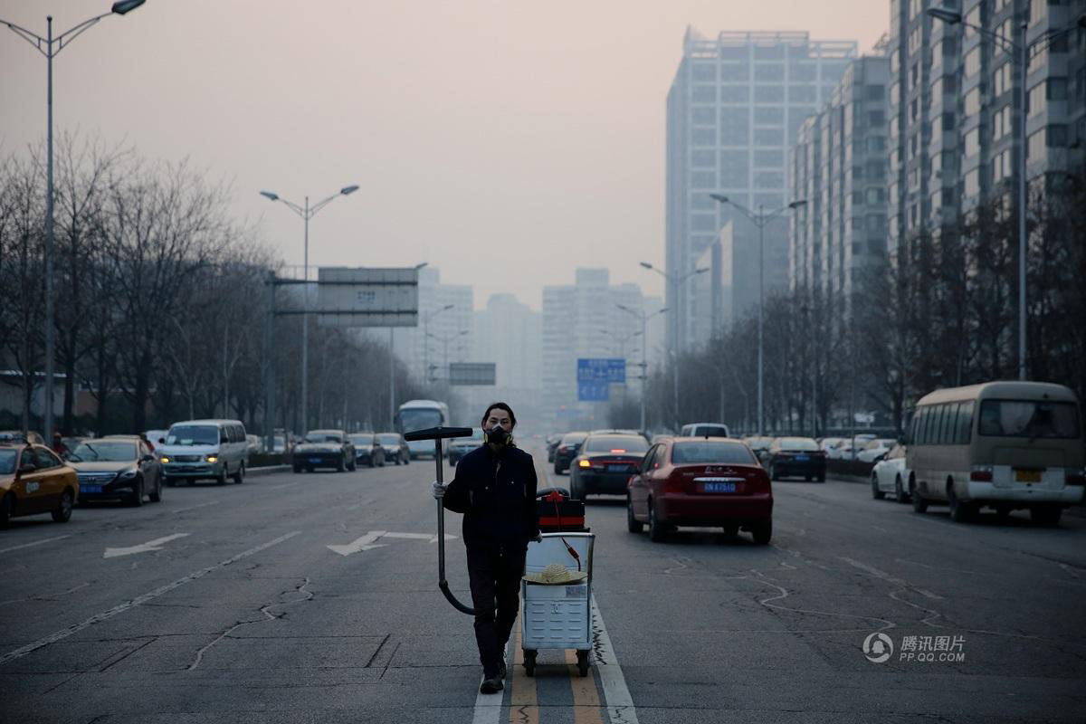 A man in a mask stands against a smoggy city skyline in China. He is pulling a vacuum cleaner down the middle of a busy street