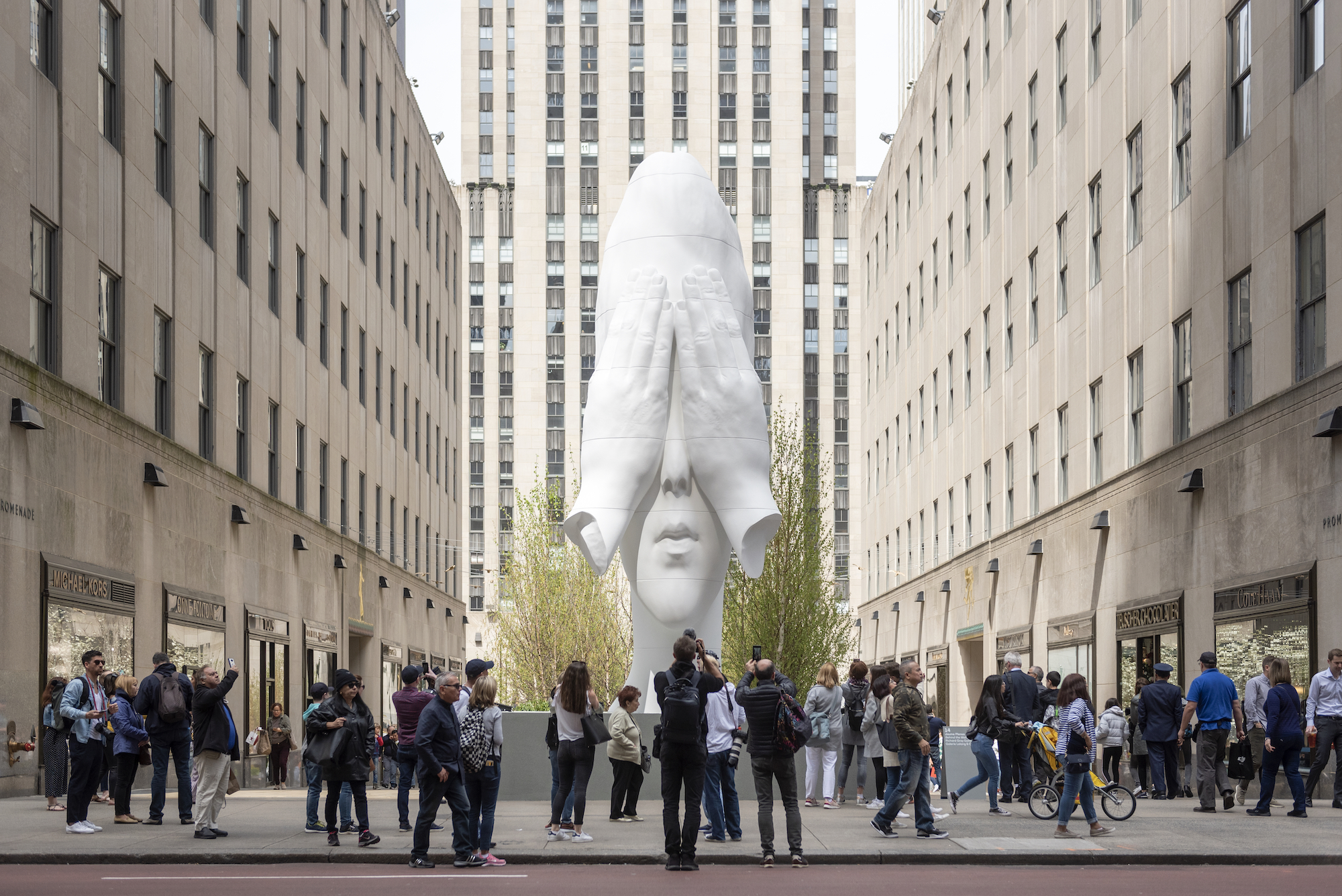 A crowd of people stand around a very tall, white sculpture depicting a human face with two hand covering it's eyes