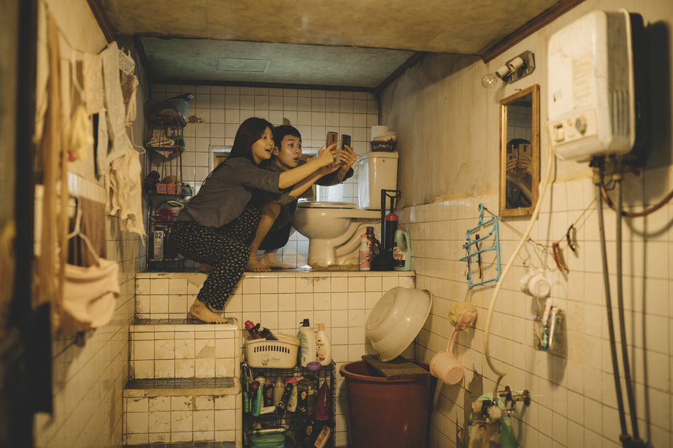 A still from the film Parasite. Two people hold phones selfie-style in a cluttered, rundown bathroom