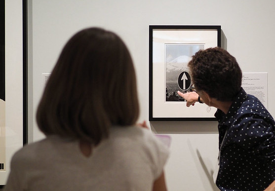 An instructor on the right points their finger toward a painting on the wall, while a student socially distances in the foreground observing the instructor.