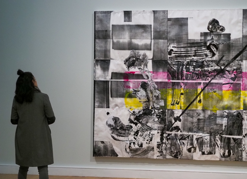 A woman stands looking at a gray, yellow, and pink painting.