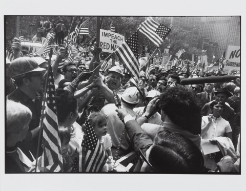 This photograph depicts a scene at a crowded political demonstration in a city. The crowd is waving flags and signs, and a reporter is extending a microphone to a yelling man wearing a construction hardhat.