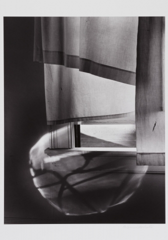 A black and white photograph showing fabric draped over a window. Light shines through the bottom half