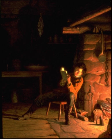 A figure wearing dark boots, dark pants, and brown overshirt reads, sitting on stool with his back to lit fireplace. Set in darkened domestic interior, table and drying herbs visible at back, wooden floorboards and stone of fireplace are illuminated. (Larson 2/5/18) 