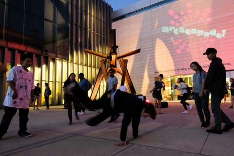 People crowd around a brown steel sculpture in the shape of a swing. One young man is mid-breakdance while others watch