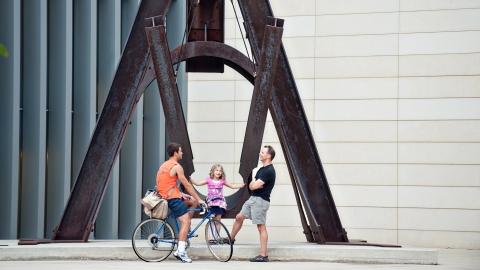 A young girl in a pink shirt sits on a large brown outdoor sculpture shaped like a swing. Two men stand in front of her talking, one is on a bicycle