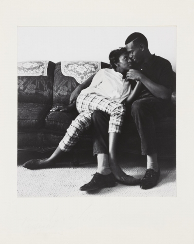 Black and white image of a man and woman sitting on a couch. The woman is leaning against the man and has one of her legs draped over one of his legs. He holds her hand. 