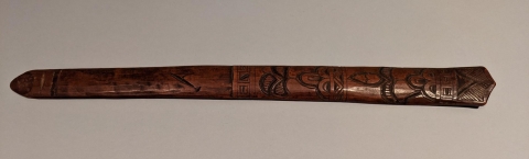 A flat wooden stick coming to a point at each end. It has oval and tear-drop shaped abstract carvings on the surface with diagonal lines at the end.