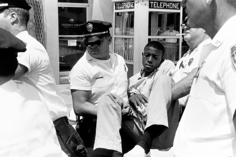 A Black teen is carried away by police officers - the faces of the police officers are naturally obscured in various ways