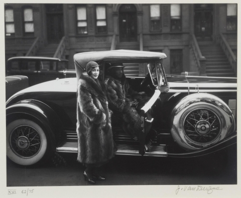 This is a photograph of an African American couple. A man sits inside his car, while a woman stands just outside, as if she is about to sit in the passenger seat. They both wear large raccoon coats. 