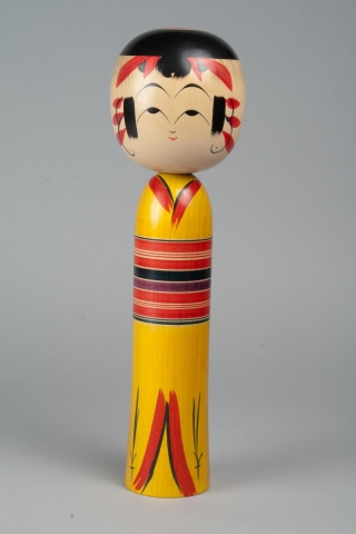 A wooden doll with two tiers made up of a head and body with no arms, legs or feet. Painted on the head is a face, hair, and a red headdress. The body is painted to look like it is wearing a red and yellow kimono with stripes red, black, and purple in the middle for the obi. You can see where the kimono begins and ends at the top and bottom of the body.