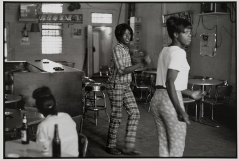 A photograph of three women in a restaurant. On the left, a woman sits at a table with glass bottles. To the right, two women dance, each wearing patterned, high-waisted trousers.