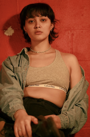 A portrait of a  seated woman. She's dressed in casual wear, a jean jacket which is opened to reveal a grey Calvin Klein Bra. She's leaning against a red wall