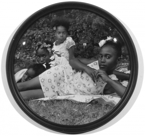 This image is a circular format black and white photograph depicting four young African American girls wearing floral dresses lounging on a blanket in the grass. Three girls sit or prop themselves up and look at the camera, the fourth girl lays down, with her eyes closed. The glass in the frame has a domed surface, creating a fish-eye effect.